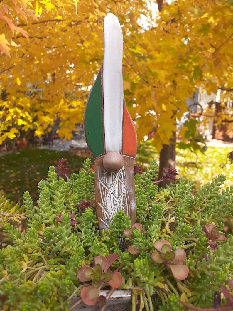 May include: A ceramic garden gnome with a white, green, and orange painted design. The gnome is standing in a pot of green succulents.