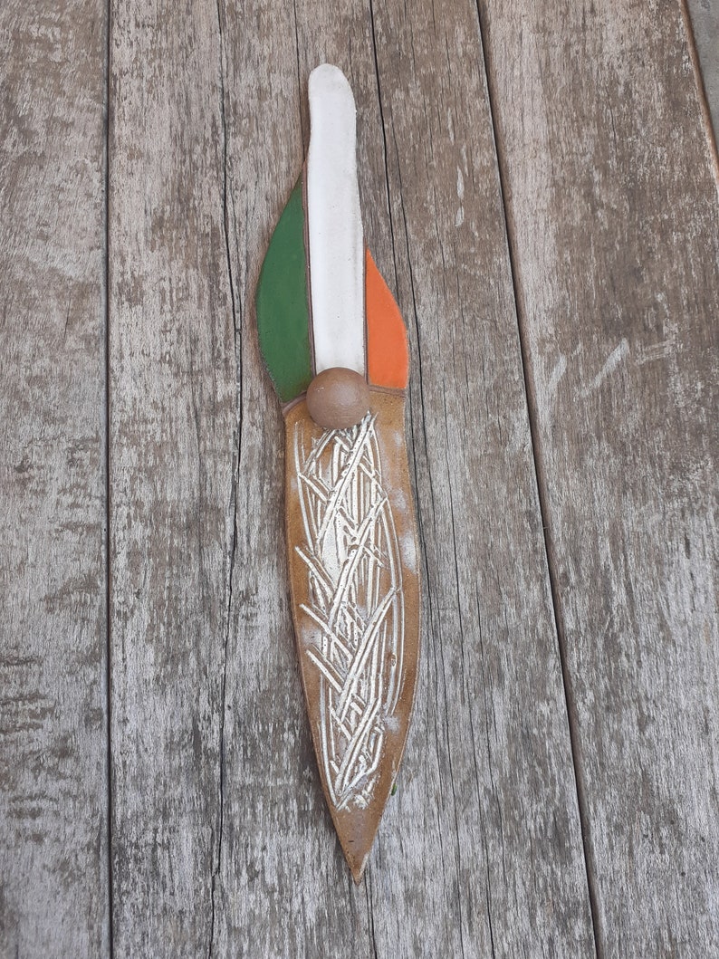 May include: A brown leather knife with a white, green, and orange design. The knife has a silver-colored design of a wheat stalk on the blade.