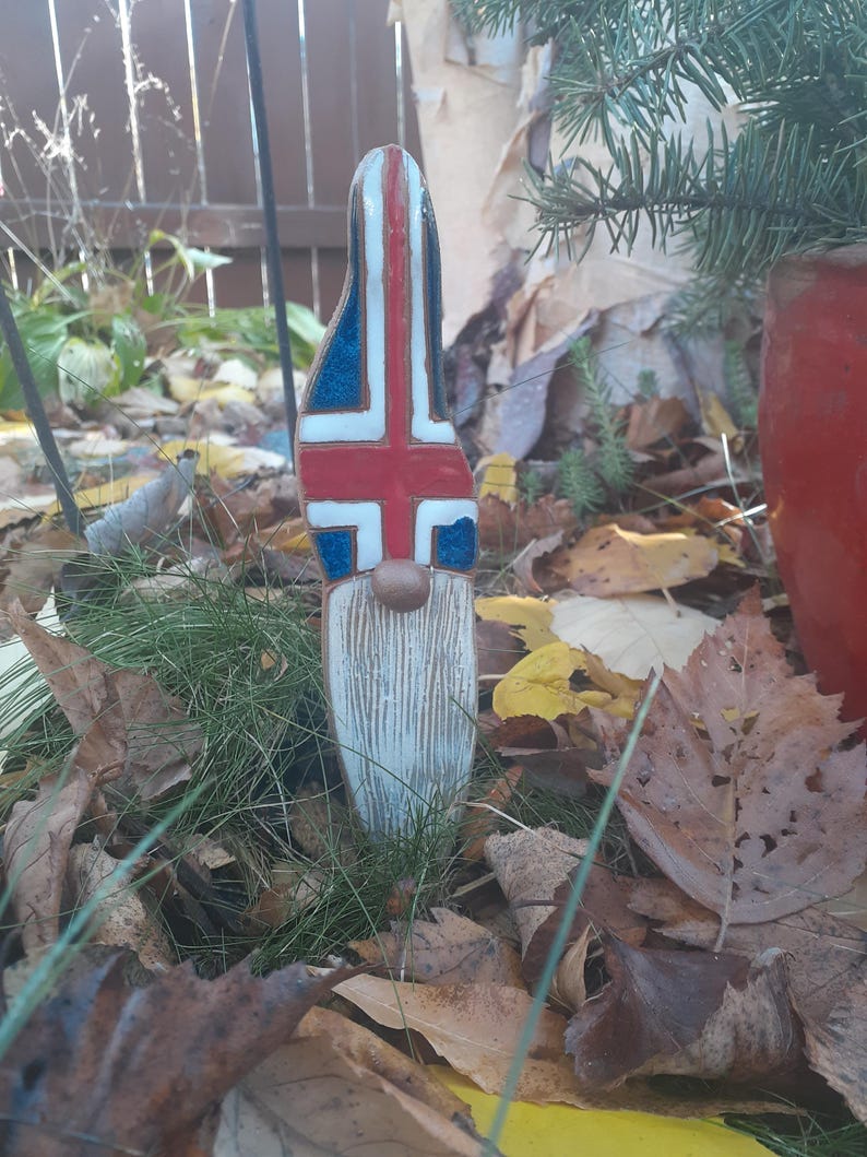 May include: A ceramic gnome figurine with a white beard and a hat painted with the Icelandic flag. The gnome is surrounded by fallen leaves and grass.