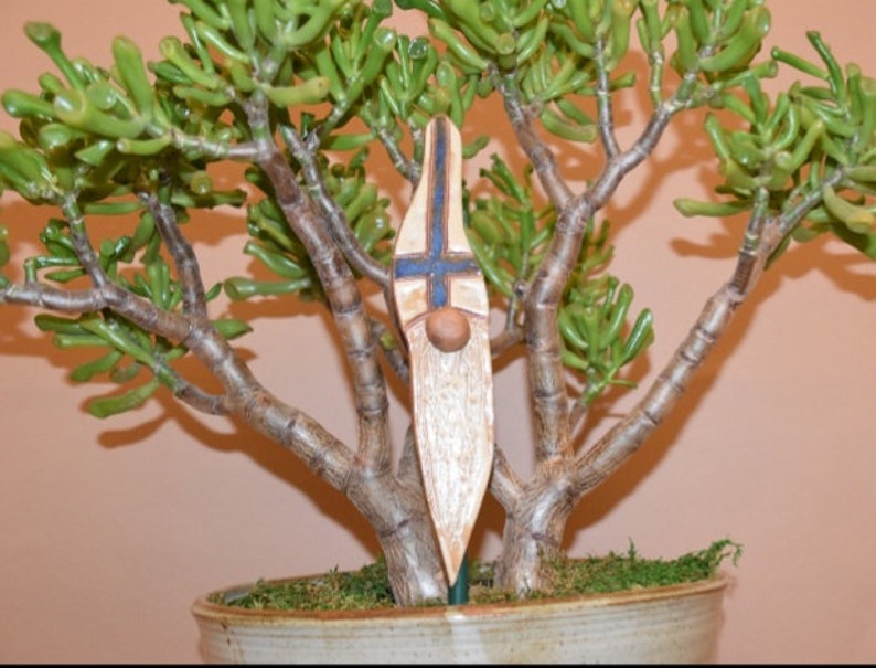 May include: A ceramic gnome figurine with a white beard and blue and white patterned hat. The gnome is standing in a pot with a green plant.
