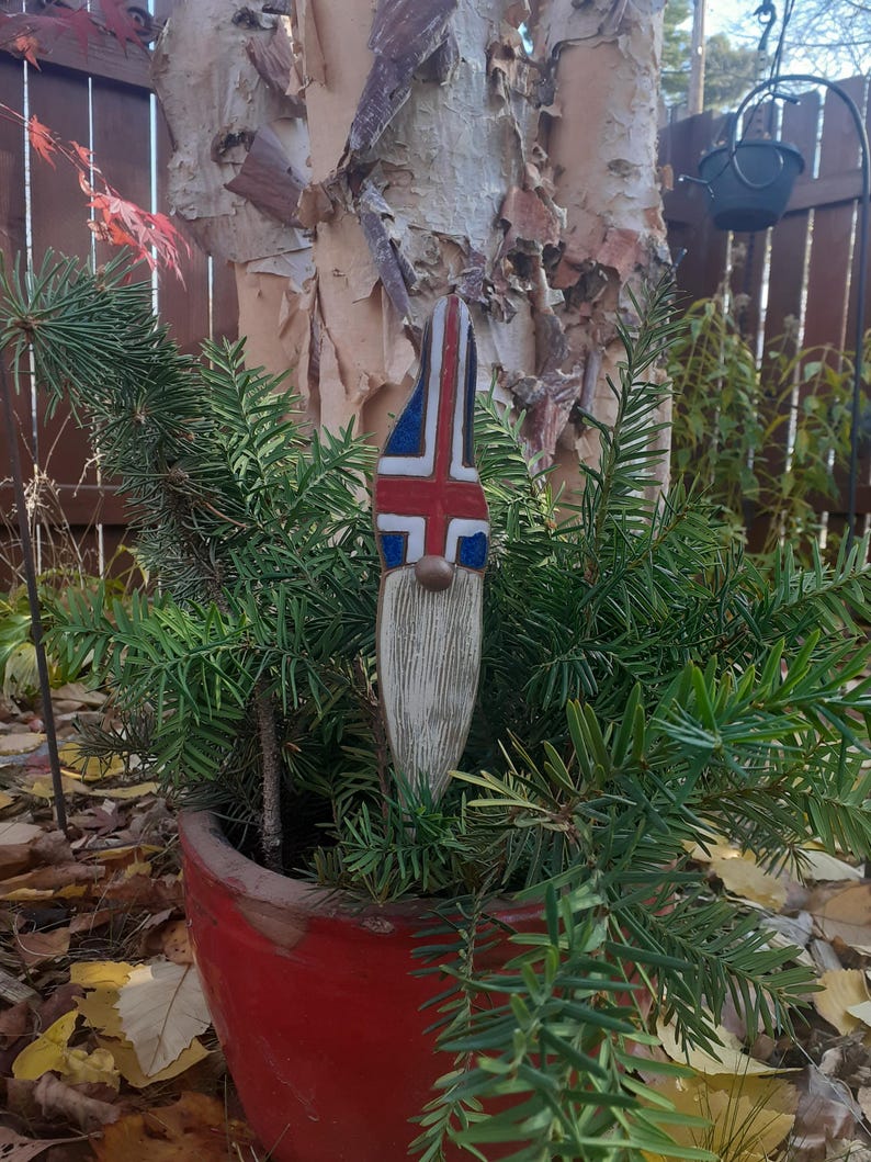 May include: A wooden gnome figurine with a white beard and a painted Icelandic flag on its hat. The gnome is standing in a red pot filled with green foliage.