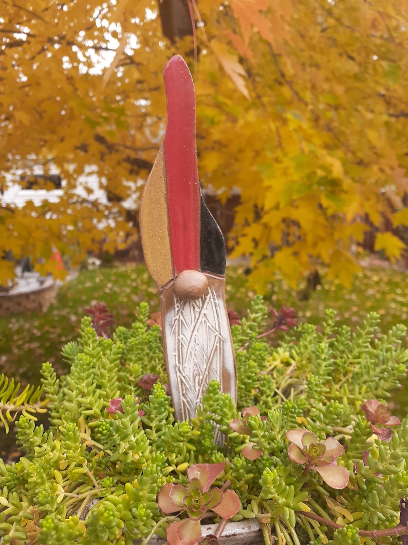 May include: A ceramic gnome figurine with a red, yellow, and black hat. The gnome is standing in a pot of green succulents.