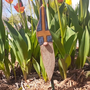 May include: A ceramic gnome figurine with a blue and yellow painted design resembling a flag. The gnome is standing in a bed of brown mulch with green plants in the background.