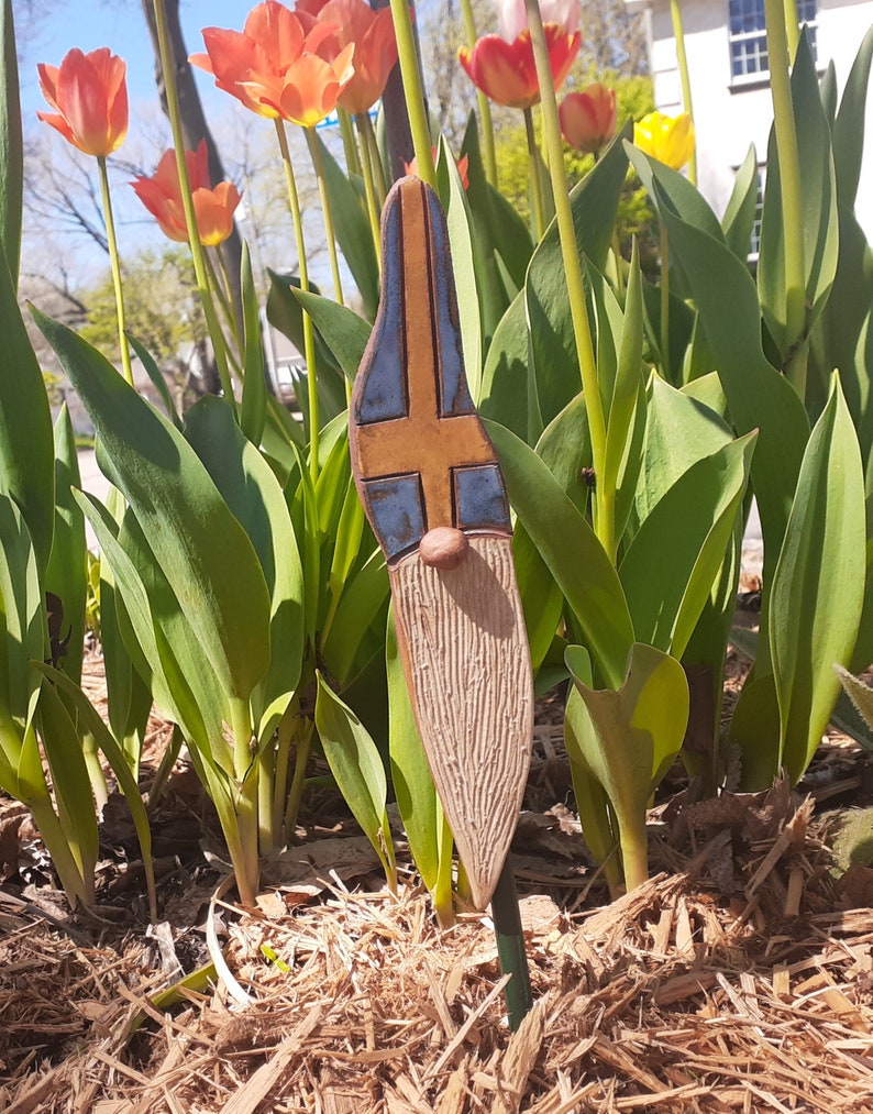 May include: A ceramic gnome figurine with a blue and yellow painted design resembling a flag. The gnome is standing in a bed of brown mulch with green plants in the background.
