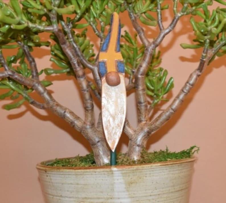 May include: A wooden gnome figurine with a painted face and a yellow, blue and white flag design. The gnome is standing in a pot with green moss and a small tree.