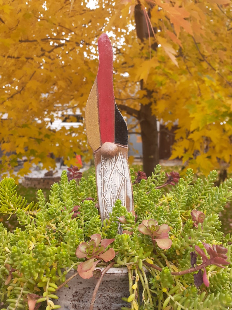May include: A ceramic gnome figurine with a red, black, and yellow hat. The gnome is standing in a pot of green succulents.