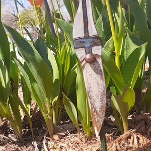 May include: A ceramic gnome garden stake with a white and blue painted design. The gnome has a brown beard and a brown hat. The stake is surrounded by green plants and brown mulch.