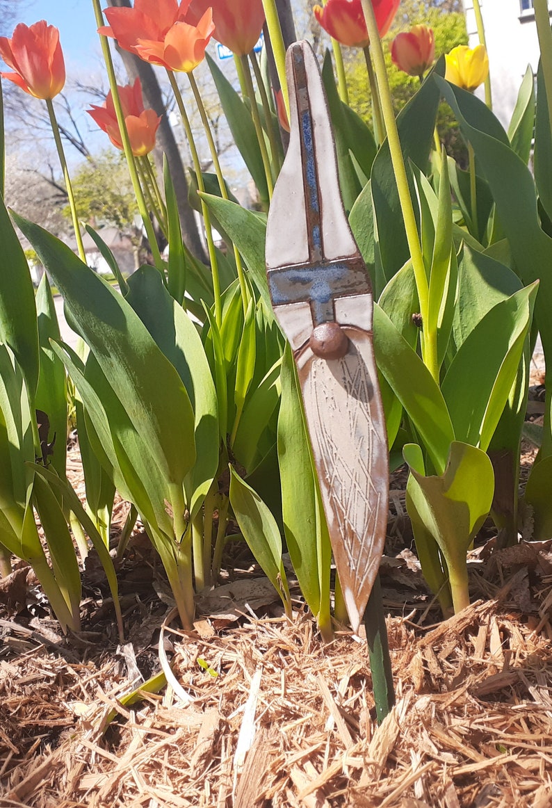 May include: A ceramic gnome garden stake with a white and blue painted design. The gnome has a brown beard and a brown hat. The stake is surrounded by green plants and brown mulch.