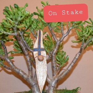 May include: A wooden gnome figurine with a blue and white cross on its chest, a brown beard, and a white painted face. The gnome is on a stake and is placed in a potted plant.