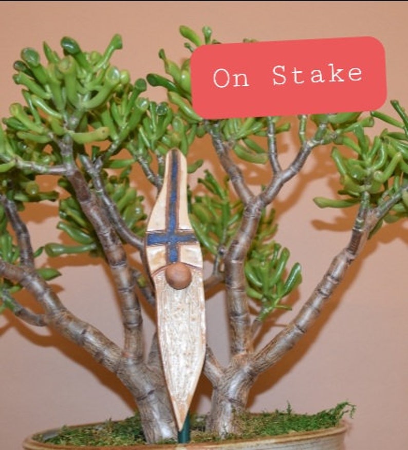 May include: A wooden gnome figurine with a blue and white cross on its chest, a brown beard, and a white painted face. The gnome is on a stake and is placed in a potted plant.