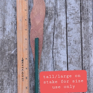 May include: A brown wooden stake with a green metal rod inserted into it. The stake is about 14 inches long. The text "tall/large on stake for size use only" is written on a red square.