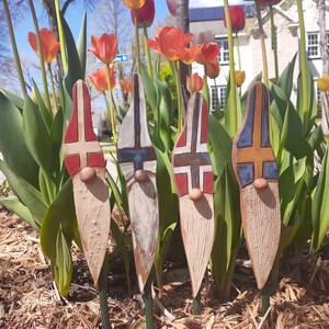 May include: Four ceramic garden gnomes with painted flags of Norway, Finland, Iceland, and Sweden. The gnomes are standing in a bed of mulch with tulips in the background.