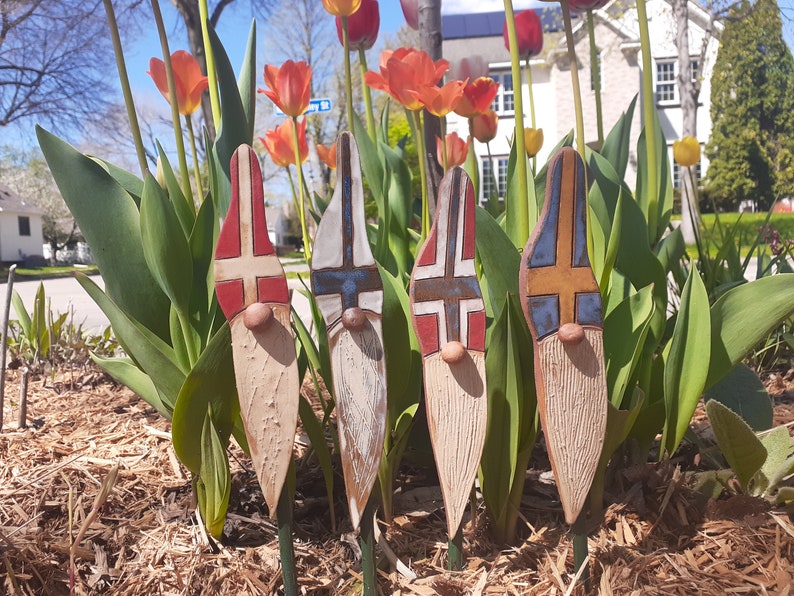May include: Four ceramic garden gnomes with painted flags of Norway, Finland, Iceland, and Sweden. The gnomes are standing in a bed of mulch with tulips in the background.