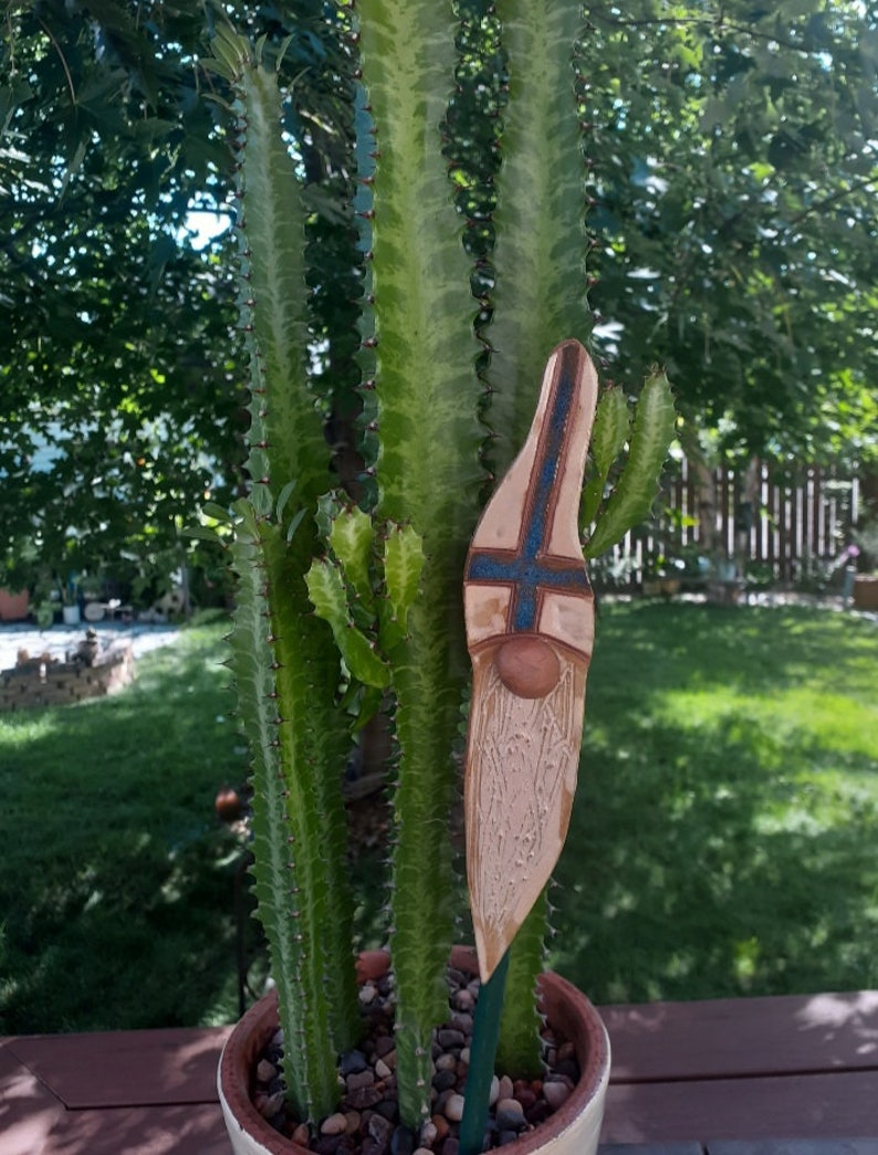 May include: A ceramic garden gnome figurine with a blue and white patterned hat and a white beard. The gnome is standing in a pot with a green cactus plant.
