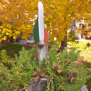 May include: A ceramic gnome figurine with a green, white, and orange hat. The gnome is standing in a pot of green succulents.