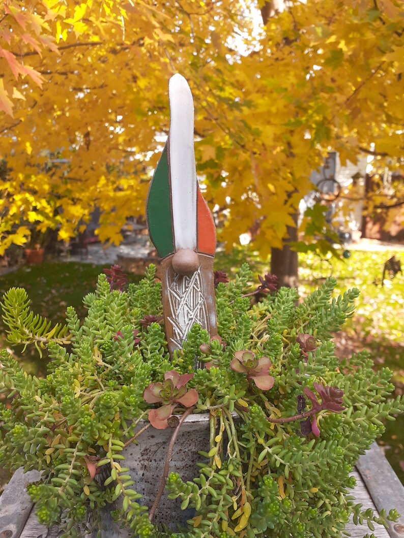May include: A ceramic gnome figurine with a green, white, and orange hat. The gnome is standing in a pot of green succulents.