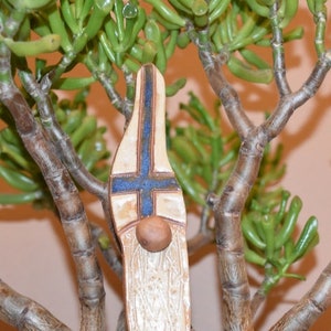 May include: A ceramic gnome figurine with a white and blue painted design. The gnome is wearing a pointed hat and has a beard. The gnome is standing in front of a green plant.