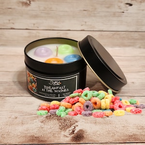 May include: A black candle tin with a white lid. The candle is labeled "Breakfast in the Rockies" and has a soy blend wax. The candle is decorated with colorful cereal rings.