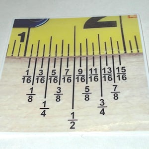 May include: A square, clear plastic ruler with a yellow tape measure design at the top. The ruler displays fractions in inches, including 1/16, 1/8, 1/4, 3/8, 1/2, 5/8, 3/4, and 7/8.