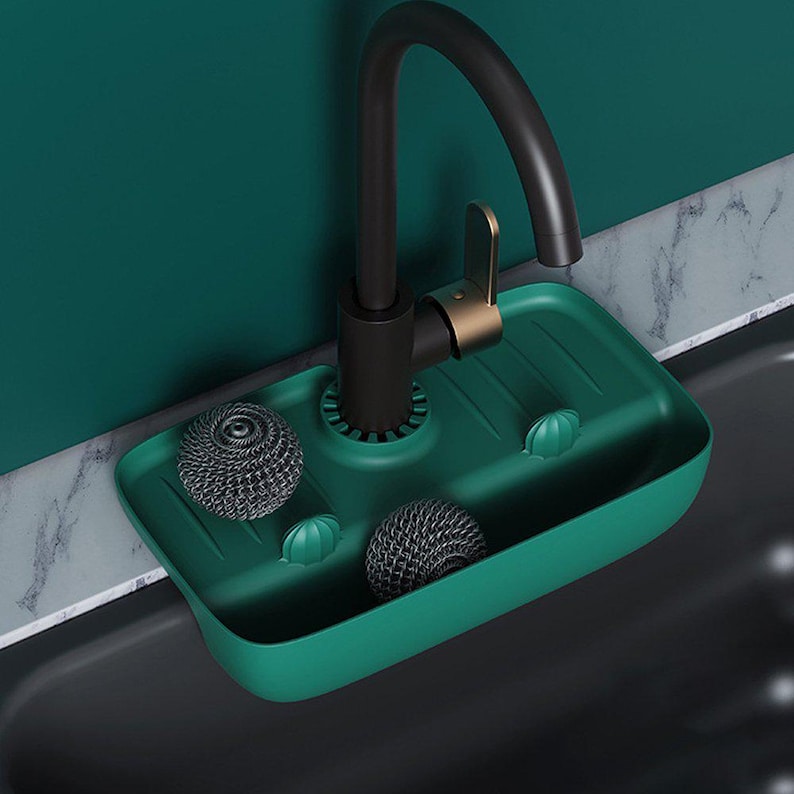 May include: A green sink caddy with a black faucet. The caddy holds two metal scrubbers and two small green holders. The faucet is black with gold accents. The sink is black, and the background is a teal color.
