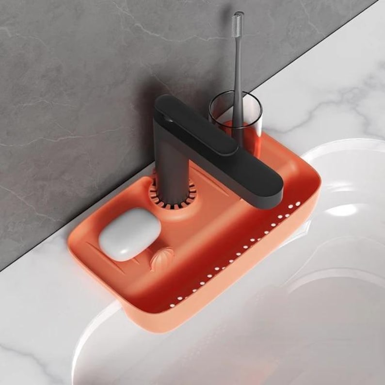 May include: An orange sink organizer with a black faucet, a bar of soap, and a toothbrush in a glass. The organizer has a rectangular shape with small holes along the edge. The faucet is black and has a modern design.