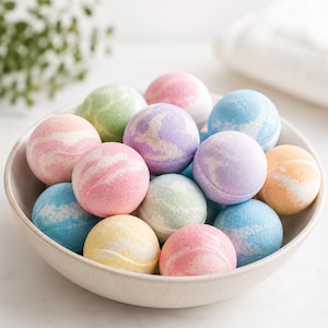 May include: A collection of spherical bath bombs in a variety of pastel colors including pink, blue, yellow, green, and purple. Each bath bomb features a distinct marbled or swirled pattern with white accents, arranged together in a shallow bowl.