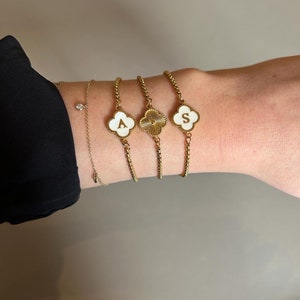 Könnte beinhalten: Drei goldene Armbänder mit weißen Kleeblatt-Anhängern. Jeder Anhänger hat einen goldenen Buchstaben, A, S, und ein Design mit einem Sonnenstrahlenmuster.