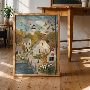 Puede incluir: Una pintura de paisaje enmarcada representa un pueblo costero con edificios blancos, un faro y una masa de agua. La obra de arte se encuentra sobre un fondo de follaje otoñal y un cielo azul. La pintura se exhibe sobre un suelo de madera.