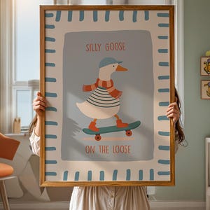 May include: A framed art print depicting a cartoon goose skateboarding. The goose is dressed in a blue cap, striped top, and scarf. The text "SILLY GOOSE ON THE LOOSE" is displayed in red and black. The frame is a light brown colour.