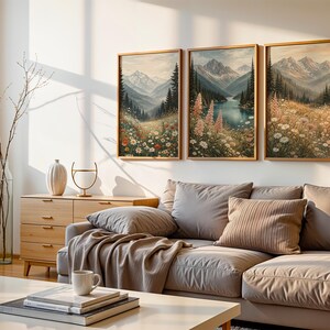 May include: Three framed landscape paintings depicting mountain scenes with colorful wildflowers and a lake. The artwork is displayed above a gray sectional sofa with throw pillows and a blanket. A wooden cabinet and coffee table are in the foreground.
