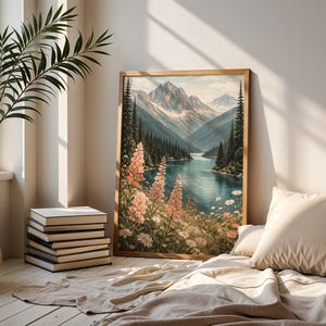 May include: A framed landscape painting depicts a serene mountain lake scene. The artwork features a wooden frame, a blue lake, snow-capped mountains, and colorful wildflowers. A stack of books and a decorative plant are in the foreground.