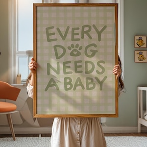 Op de afbeelding: Een ingelijste print met de tekst "EVERY DOG NEEDS A BABY" in groene letters. De achtergrond heeft een lichtgroen ruitjespatroon. De lijst is van hout. De print wordt voor een persoon gehouden.