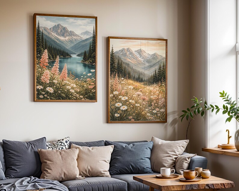 May include: Two framed landscape paintings depict mountain scenes with a lake and wildflowers. The artwork features a palette of blues, greens, and pinks, with a wooden frame. The paintings are displayed above a gray sofa with pillows.