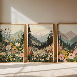 May include: Three framed landscape art prints. Each print features a mountain scene with colorful wildflowers in the foreground. The frames are gold-colored, and the artwork has a vintage aesthetic. The prints are displayed on a light-colored surface.