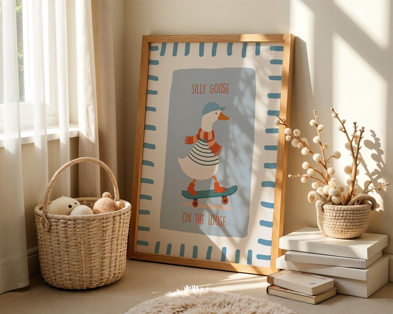 May include: A framed art print with a cartoon goose on a skateboard, wearing a hat and scarf. The print reads "SILLY GOOSE ON THE LOOSE" and has a light blue background with a light brown frame. A woven basket and decorative items are in the foreground.