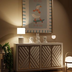 May include: A framed print with a cartoon goose on a skateboard, with the text "SILLY GOOSE ON THE LOOSE." The print is above a white cabinet with gold detailing, and decorative animal figurines. A white lamp and a potted plant are also visible.