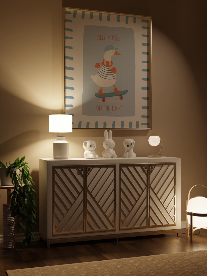 May include: A framed print with a cartoon goose on a skateboard, with the text "SILLY GOOSE ON THE LOOSE." The print is above a white cabinet with gold detailing, and decorative animal figurines. A white lamp and a potted plant are also visible.