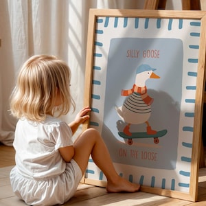 May include: A framed art print with a cartoon goose on a skateboard, wearing a blue cap and scarf. The print has a light blue background with the words "Silly Goose On The Loose" in orange text. The frame is made of light wood.