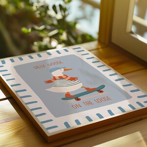 May include: A framed art print with a cartoon goose on a skateboard. The goose is wearing a blue cap, striped shirt, and scarf. The text reads "Silly Goose On The Loose" in orange. The frame has a light blue border.