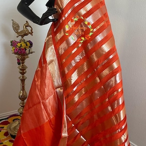 Peut inclure: Un sari orange vif avec des rayures et des bordures dorées est drapé sur un mannequin noir. Le tissu a un aspect brillant, semblable à de la soie. Le sari est présenté dans un cadre bien éclairé, mettant en valeur sa couleur et son design.
