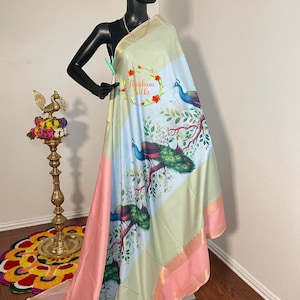 May include: A light green and pink silk saree with a white border and a peacock design. The saree has a gold border and the text "Arichcham Silks" is printed on the fabric.