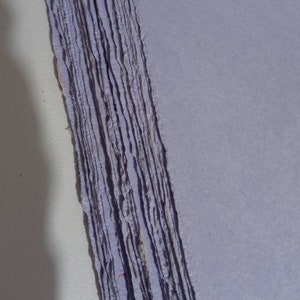 Può includere: Un mazzo di fogli di carta fatti a mano di colore lilla chiaro. La carta ha una superficie ruvida e testurizzata e bordi irregolari.