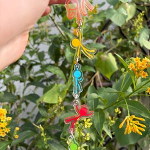 Pode incluir: Um porta-chaves vertical em acrílico com cinco figuras de pau translúcidas e coloridas em várias poses, ligadas por anéis de metal prateado. As figuras são cor de laranja, amarelas, azuis, vermelhas e verdes, cada uma numa forma de acrílico transparente.