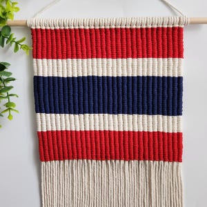 May include: A red, white and blue striped macrame wall hanging with a fringe border. The hanging is made with cotton cord and is attached to a wooden dowel.