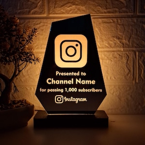 May include: A black acrylic award plaque with a gold Instagram logo and the text "Presented to Channel Name for passing 1,000 subscribers Instagram".