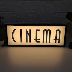 Peut inclure: Un caisson lumineux rectangulaire avec le mot "CINEMA" en lettres noires stylisées. Les lettres sont éclairées sur un fond crème. Le caisson lumineux a un cadre noir et est probablement utilisé à des fins décoratives.