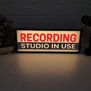 以下が含まれることがあります： 赤と白の文字で「RECORDING STUDIO IN USE」と書かれた長方形の黒い看板。看板は点灯しており、電源が入っていることを示唆しています。背景は暗いレンガの壁です。