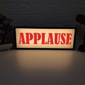以下が含まれることがあります： 白い背景に赤い文字で「APPLAUSE」と書かれた、黒い枠のライトアップされた看板です。