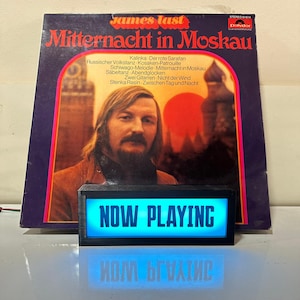 Puede incluir: Un álbum de vinilo vintage titulado "Mitternacht in Moskau" de James Last. La portada del álbum presenta un retrato de un hombre con pelo largo y bigote. Un letrero rectangular negro con las palabras "NOW PLAYING" en luz azul brillante está frente al álbum.