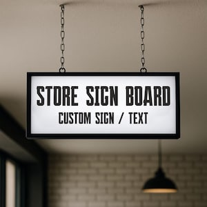 Può includere: Un'insegna rettangolare per negozi con la scritta "STORE SIGN BOARD" e "CUSTOM SIGN / TEXT" in grassetto nero. L'insegna è sospesa al soffitto da due catene di metallo. L'insegna ha una cornice nera e uno sfondo bianco illuminato.